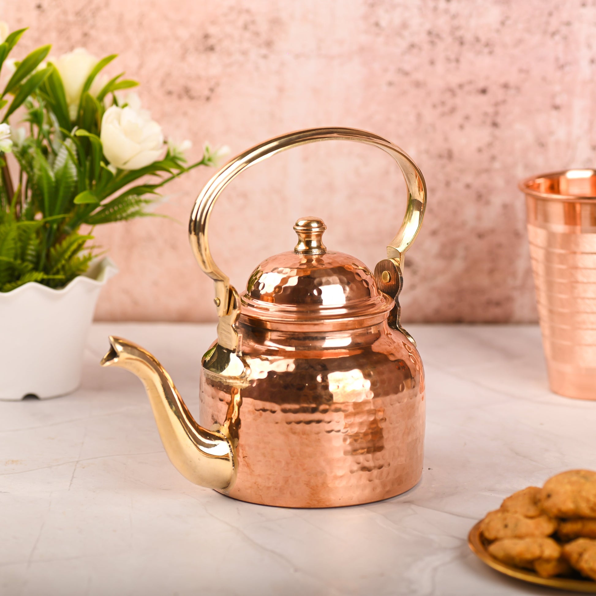 Copper teapot with gold accents on a light surface with flowers and cookies in the background - Eco-friendly - Sama Homes