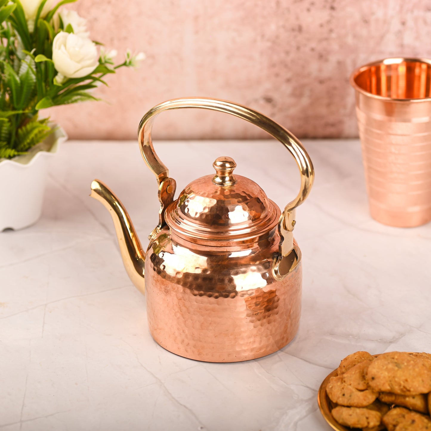 Copper teapot with gold handle on a marble surface with cookies and a cup in the background.