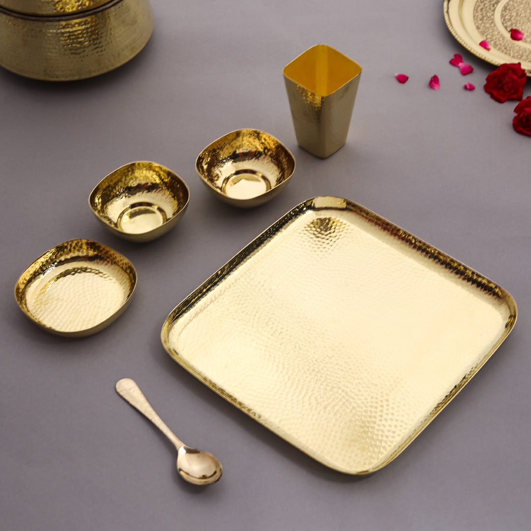 Set of gold dinnerware including a square tray, bowls, and a spoon on a gray background.
