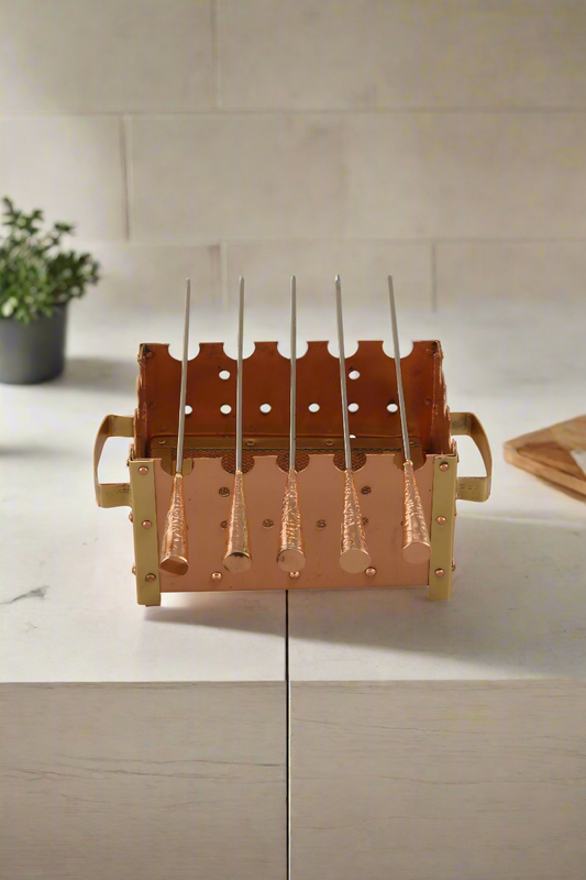 A bronze-colored barbeque grill with multiple grilling areas, situated on a patio with a white wall and greenery in the background.